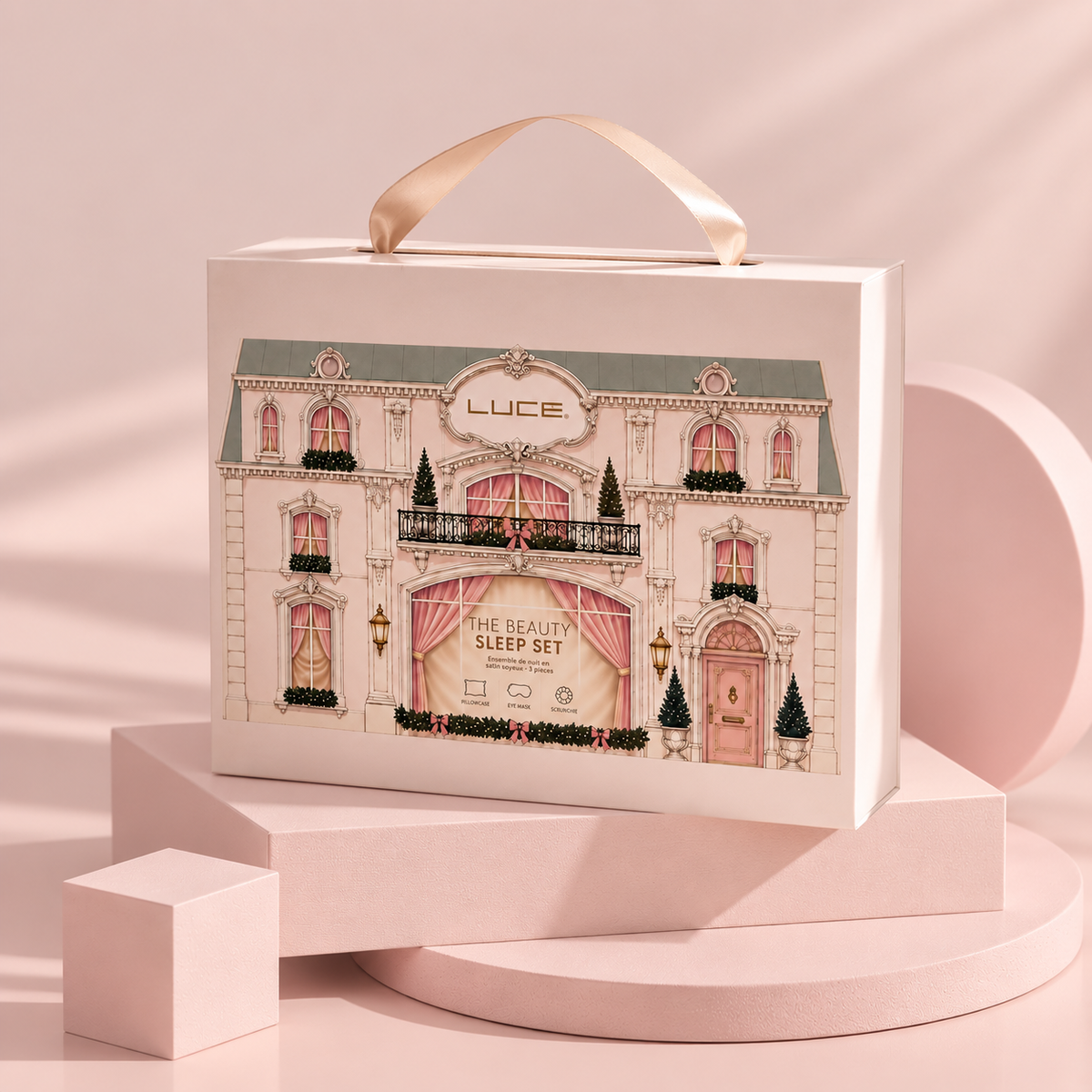 A rectangular cosmetic gift box with a handle, printed with an illustrated elegant building facade and the text 'LUCÉ' and 'THE BEAUTY SLEEP SET.' The box is photographed on pink display blocks against a soft pink studio background.