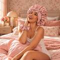 A woman smiling on a bed wearing a satin pink bonnet and matching pink scrunchie, with pink bedding and floral wallpaper in a cozy bedroom setting.